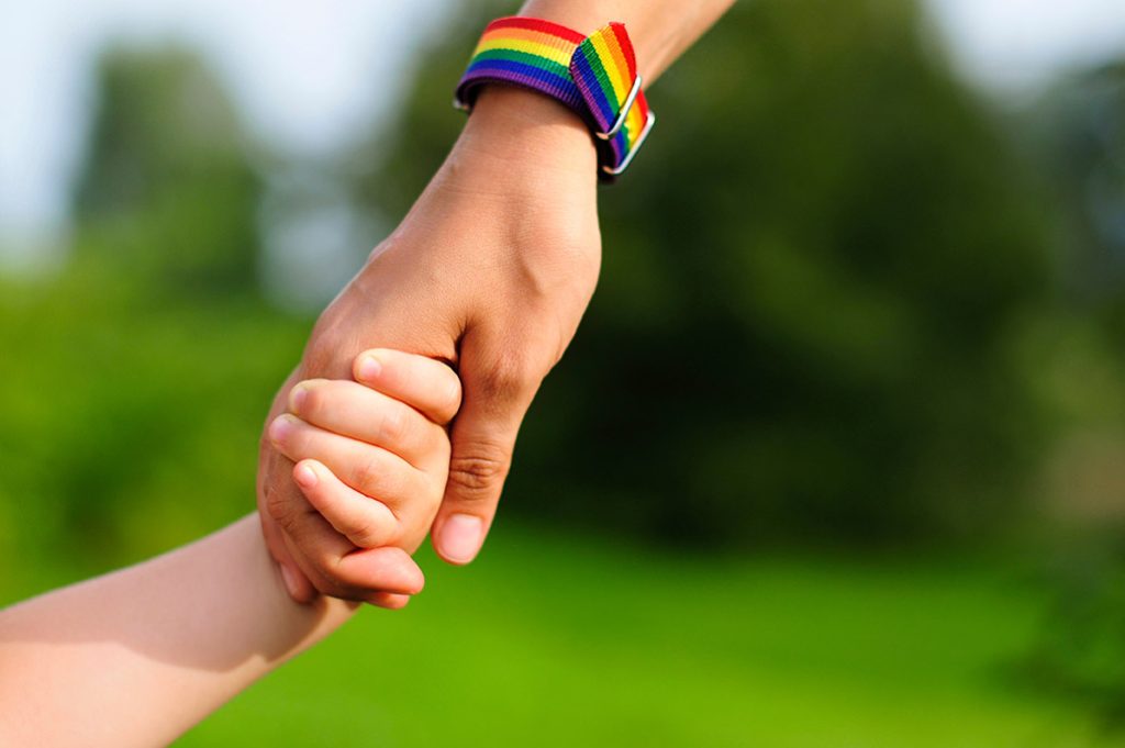 Online therapist for LGBTQ+ parents in Texas An adult hand with LGBTQ flag watch holds the hand of their child representing LGBTQ parenting