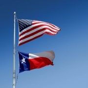 U.S. and Texas flags fly on a pole surrounded by blue skies